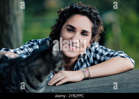 Giovane donna con capelli ricci, con una camicia a scacchi, sorride alla macchina fotografica mentre accarezza un gatto da tavola all'aperto, appoggiandosi su una superficie di legno Foto Stock