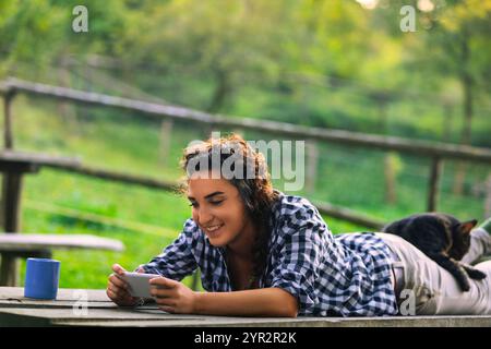 Una giovane donna giace su un tavolo in campagna. Sorridendo mentre usa il suo telefono. Con il suo gatto che dorme sulla schiena. Circondato dal verde. Le piace Foto Stock