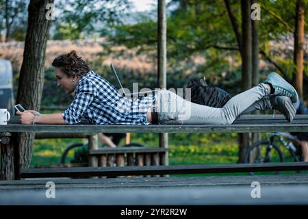 Un giovane ventenne si rilassa sulla panchina di un parco con un gatto, utilizzando il suo portatile e lo smartphone all'aperto. Godersi la natura mentre si lavora in remoto, emb Foto Stock