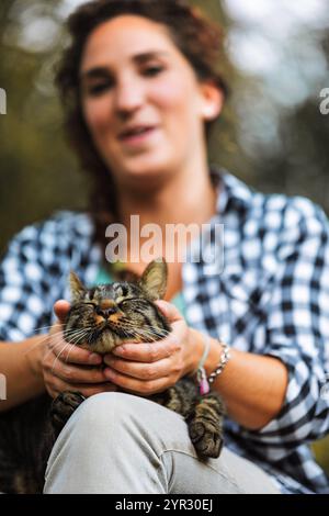 Una giovane donna tiene delicatamente il viso di un gatto da tabby all'aperto, mostrando amore e connessione. Sorridendo, condividono un momento tenero in natura, riflettendo un forte Foto Stock