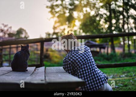 L'agricoltore e il suo gatto sono seduti su un tavolo di legno, godendosi il tranquillo tramonto in una fattoria, creando una scena emozionante di compagnia e tra rurale Foto Stock