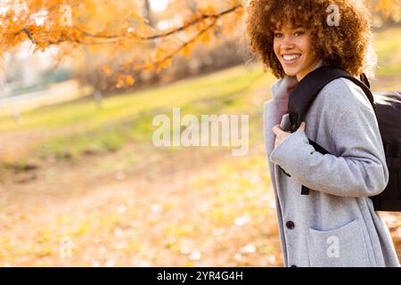 In uno splendido parco autunnale, una giovane donna gioiosa dai capelli ricci mostra il suo sorriso brillante indossando un elegante cappotto grigio e un maglione rosso, carryin Foto Stock