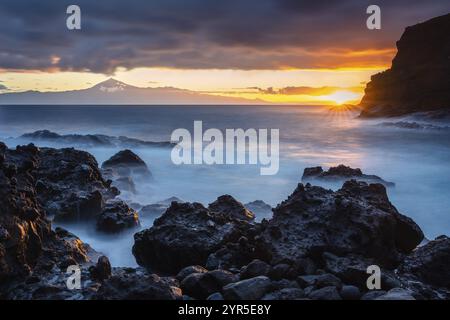 Alba sulla spiaggia di San Marcos. Rocce, mare e sole. Tenerife e il Teide sullo sfondo. Esposizione lunga. La Gomera, Isole Canarie, Spagna Foto Stock