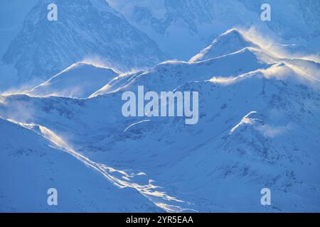 Cime innevate retroilluminate da neve volante, su un cielo azzurro limpido in un paesaggio ventoso, Leukerbad, Leuk, Vallese, Svizzera, Europa Foto Stock