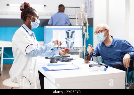 Paziente in pensione caucasica in consultazione con un medico donna, discutendo i risultati dei raggi x. Il medico afroamericano mostra l'immagine della radiografia polmonare all'uomo anziano in sedia a rotelle e spiega la diagnosi. Foto Stock