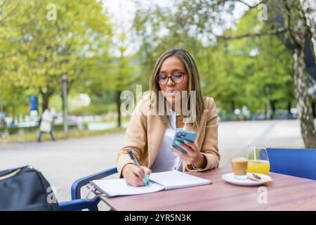 Donna d'affari latina che scrive appunti mentre lavora utilizzando il telefono cellulare seduto in una caffetteria sul marciapiede Foto Stock