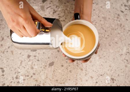 Una persona versa il latte montato in una tazza da caffè nera, creando un latte con una macchina per espresso sullo sfondo. La schiuma di latte si fonde con Foto Stock