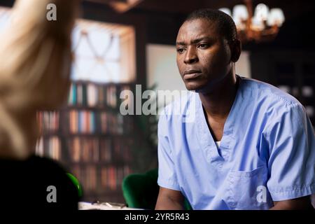 Professionista sanitario nero in scrub durante una sessione di consulenza, discutendo il bilancio emotivo della cura del paziente. Infermiera maschile condivide le sue lotte come personale medico che lavora in ospedale. Foto Stock