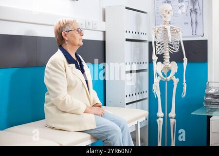 Donna anziana vestita casualmente siede in una stanza d'ospedale preparata per la sua consultazione sanitaria. Anziana signora caucasica aspetta sul letto d'esame per il controllo medico in ufficio medici. Foto Stock