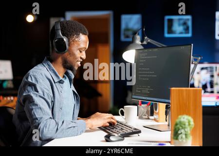 Sviluppatore web afroamericano che codifica script server su pc, utilizzando codice binario e linguaggio di programmazione sulla finestra terminale. Il debug della protezione del database da parte di un amministratore di sistema giovane. Foto Stock