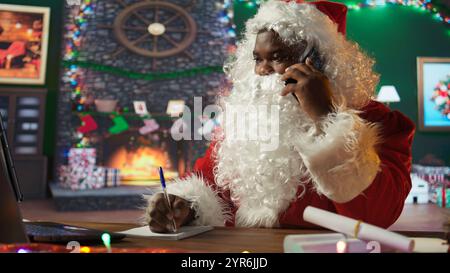 San Nicola afroamericano chiama genitori e bambini su smartphone, preparandosi per il viaggio durante le vacanze della vigilia di Natale. L'uomo con la barba bianca porta giocattoli e regali. Telecamera B.. Foto Stock