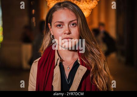 Giovane donna seria con lunghi capelli biondi e una sciarpa rossa, in piedi di notte sotto le luci festose, che guarda direttamente la fotocamera con un'espressione mirata, Lady con un atteggiamento pensivo che guarda davanti. Foto di alta qualità Foto Stock