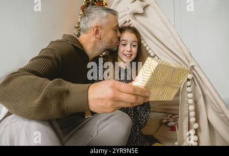 Un momento di Natale emozionante: Un padre condivide un tenero bacio con sua figlia mentre le regala un regalo all'interno della loro accogliente tepee. La scena trasuda hol Foto Stock