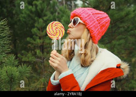 Ritratto invernale di una giovane donna felice divertente sorridente che si diverte con un dolce lecca lecca-lecca che indossa un cappello contro l'albero di Natale nella foresta Foto Stock