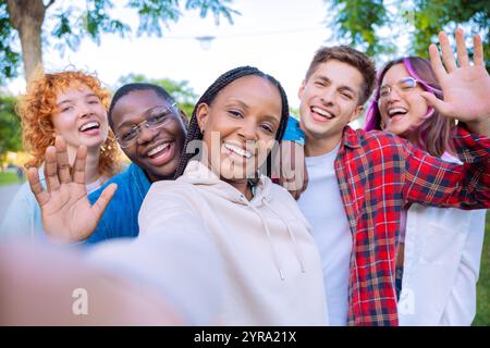 Felice gruppo di amici diversi che scattano un selfie all'aperto, sorridono e salutano Foto Stock