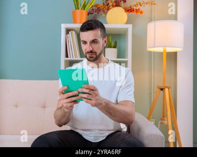 Uomo seduto su un divano, che usa un tablet in un salotto accogliente e moderno Foto Stock