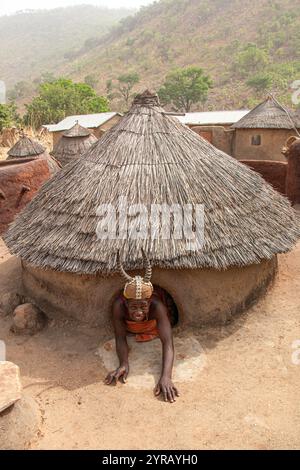 Donna in costume tradizionale con cappello cornuto che emerge da una casa di argilla in un villaggio del Togo Foto Stock