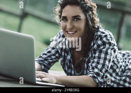 Giovane donna con capelli ricci. Indossa una camicia a scacchi. Giace su una superficie di legno all'aperto. Lavora diligentemente sul suo notebook. Goditi l'aria fresca Foto Stock