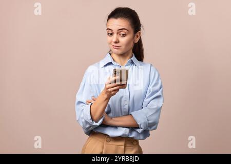 donna scettica in camicia blu che tiene in mano il cellulare che guarda lo smartphone su sfondo beige. Foto Stock