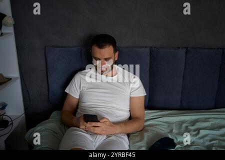 un uomo di 30 anni con macchie idratanti sotto gli occhi sta chiacchierando su uno smartphone Foto Stock