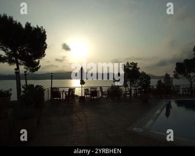 Una vista serena di un tramonto che proietta sfumature dorate sulle acque calme, con sagome di alberi e posti a sedere all'aperto. L'orizzonte presenta montagne lontane, Foto Stock