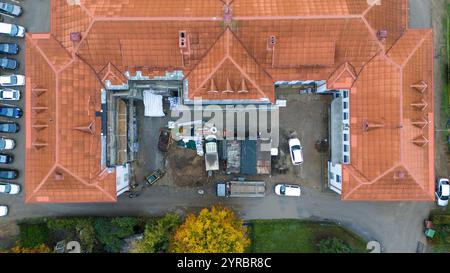 Vista aerea di un cantiere edile su un edificio con tetti di tegole rosse, circondato da auto parcheggiate e vegetazione. Foto Stock