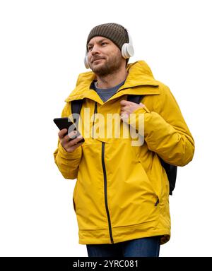 Uomo caucasico con giacca e cappello invernali gialli, in piedi con un telefono cellulare, cuffie wireless e uno zaino. Foto Stock