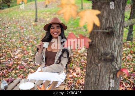 La giovane donna latina sorridente che indossa un cappello si gode una tazza di caffè a un tavolo di legno in un colorato parco autunnale, circondato da foglie e alberi caduti Foto Stock