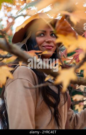 La giovane donna latina con lunghi capelli scuri e un cappello marrone sorride serenamente mentre si trova dietro le foglie dorate di un ramo d'albero in autunno Foto Stock