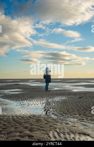 Donna in piedi su una spiaggia vuota con la bassa marea e indossa un parka invernale Foto Stock