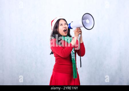 ritratto di donne indonesiane che indossano maglione natalizio, cappello e sciarpa di babbo natale, urlando arrabbiato in alto a sinistra, schiava entrambe le mani con il megafono, Foto Stock