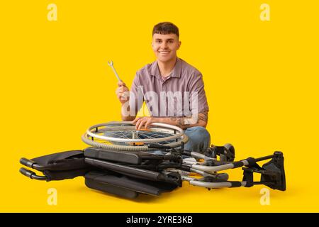 Bel giovane uomo felice con chiave che ripara sedia a rotelle su sfondo giallo Foto Stock