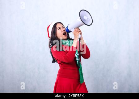 espressione di donne asiatiche adulte che indossano un maglione natalizio rosso con cappello di babbo natale e sciarpa, urlando molto arrabbiata in alto a sinistra, entrambe le mani che tengono il megafono Foto Stock