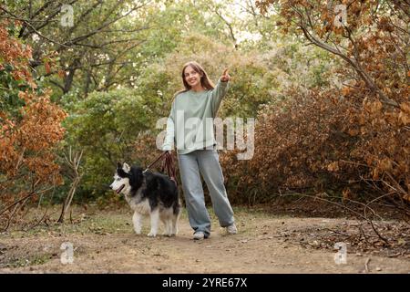 Bella giovane donna con un simpatico cane Husky che indica qualcosa e cammina nel parco Foto Stock