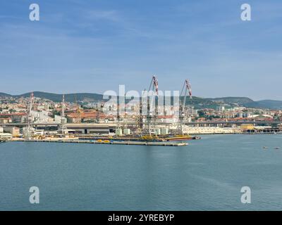 Trieste, Italia - 29 giugno 2024: Porto commerciale. Grosse gru sui moli senza navi. Paesaggio urbano e catena montuosa all'orizzonte Foto Stock