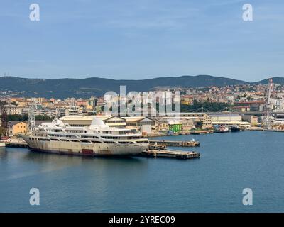 Trieste, Italia - 29 giugno 2024: Porto commerciale. Vecchia nave passeggeri fuori Kingstown attraccata, con paesaggio urbano e catena montuosa all'orizzonte Foto Stock