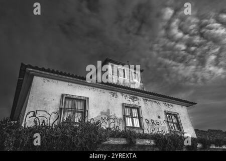 La facciata sbriciolante di una casa abbandonata presenta graffiti e finestre sbarrate, adagiata su un cielo spettacolare e nuvoloso in monocromia Foto Stock