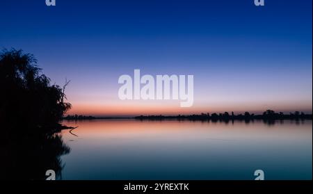 I colori vivaci del tramonto si riflettono sul lago calmo, creando una scena tranquilla con sagome di alberi che aggiungono tranquillità Foto Stock