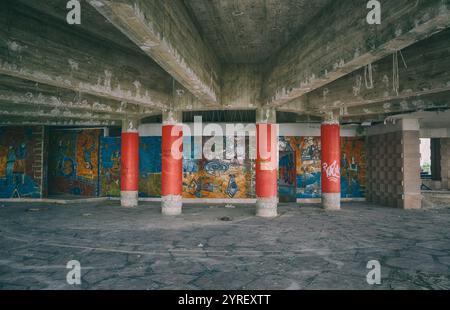 Colonne di cemento che sostengono un soffitto decadente si stagliano in un edificio abbandonato, mostrando colorate opere d'arte a mosaico e graffiti sulle pareti, testimonianza di p Foto Stock