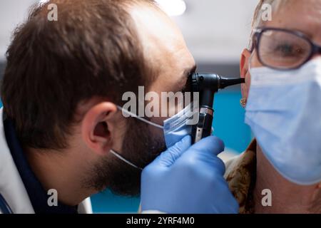 Primo piano dello specialista della ent maschile con l'otoscopio esaminando l'orecchio della donna anziana, entrambi indossando la maschera in clinica. Medico barbato che utilizza uno strumento di otologia per verificare la presenza di infezioni su pazienti in pensione. Foto Stock