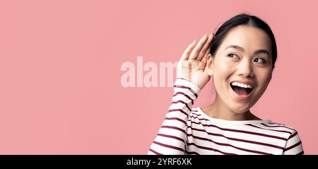 Curiosa donna asiatica allegra che tiene la mano vicino all'orecchio cercando di sentire qualcosa Foto Stock