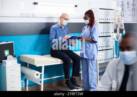 Assistente medico femminile aiuta il paziente anziano caucasico a compilare le sue informazioni mediche sugli appunti. L'uomo bianco in pensione riceve la guida da infermiera entrambi indossando la maschera in ufficio medici. Foto Stock