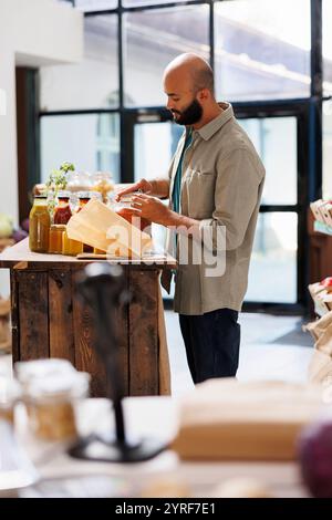 L'uomo arabo chiude il coperchio di un vaso di vetro contenente prodotti alimentari biologici. Accanto a lui sul tavolo c'è una borsa marrone. Si trova in un negozio di alimentari sano ed ecologico. Foto Stock