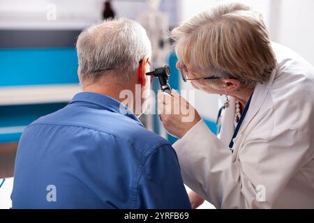 Paziente pensionato maschio in camera d'ospedale ha il suo orecchio esaminato da medico esperto. Medico donna in pensione utilizzando otoscopio su vecchio uomo bianco, assicurando diagnosi professionale in studio medico. Foto Stock