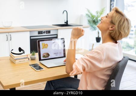 Donna caucasica sui trent'anni che ordina cibo online con un portatile, esprimendo gioia. Confortevole ambiente di cucina casalinga con arredamento accogliente. Uso di tecnologie moderne e convenienza per la consegna di alimenti. Foto Stock