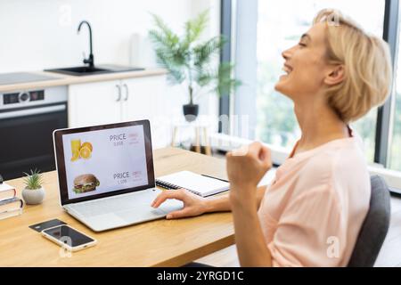 Donna caucasica felicemente ordinare cibo online utilizzando un computer portatile. Si trova in un ambiente moderno, dove prova gioia e soddisfazione con consegna di cibo scontata. Foto Stock