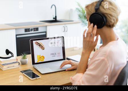 Donna caucasica felice che si diverte a fare acquisti online sul sito Web di consegna utilizzando un computer portatile mentre ascolta musica con le cuffie. Ambiente rilassato con moderno sfondo cucina. Foto Stock