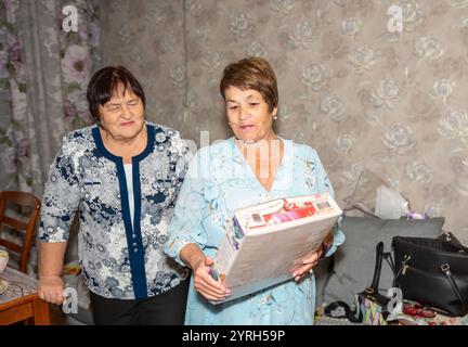 Due amici di 70 anni celebrano un'occasione speciale a casa. Uno è felicemente sorpreso da un regalo dall'altro. Entrambi sono vestiti e condividono una gioia Foto Stock