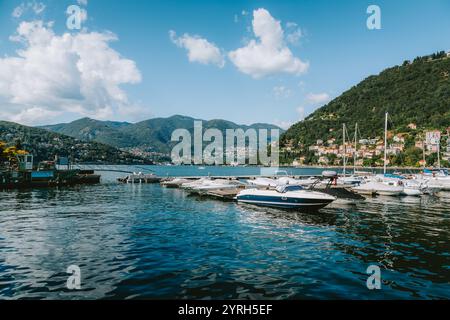 Il lago di como risplende sotto un vivace cielo blu, con numerosi motoscafi e barche a vela ormeggiati lungo la costa, sostenuti da lussureggianti colline verdi e charmin Foto Stock