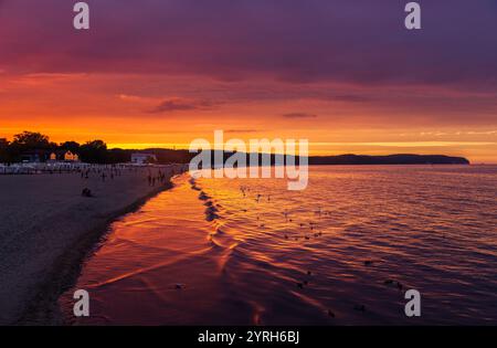 I turisti passeggiano al tramonto sulla spiaggia di sopot, con cigni e anatre che nuotano nel mare sotto un cielo colorato. Una serena serata estiva in riva al mare Foto Stock